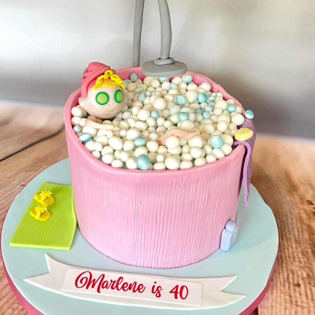 Pink bathtub cake. A lady in the bath, with lots of little bubbles. Decorated with slippers, a bathmat, shampoo and a towel.